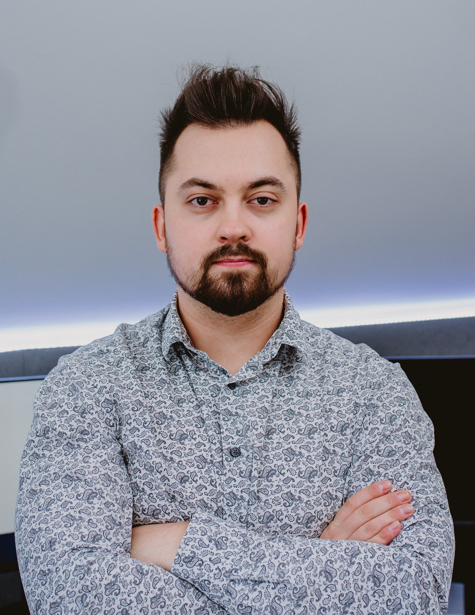 Portret mężczyzny z brodą w koszuli w paisley, z założonymi rękami, patrzącego w obiektyw.