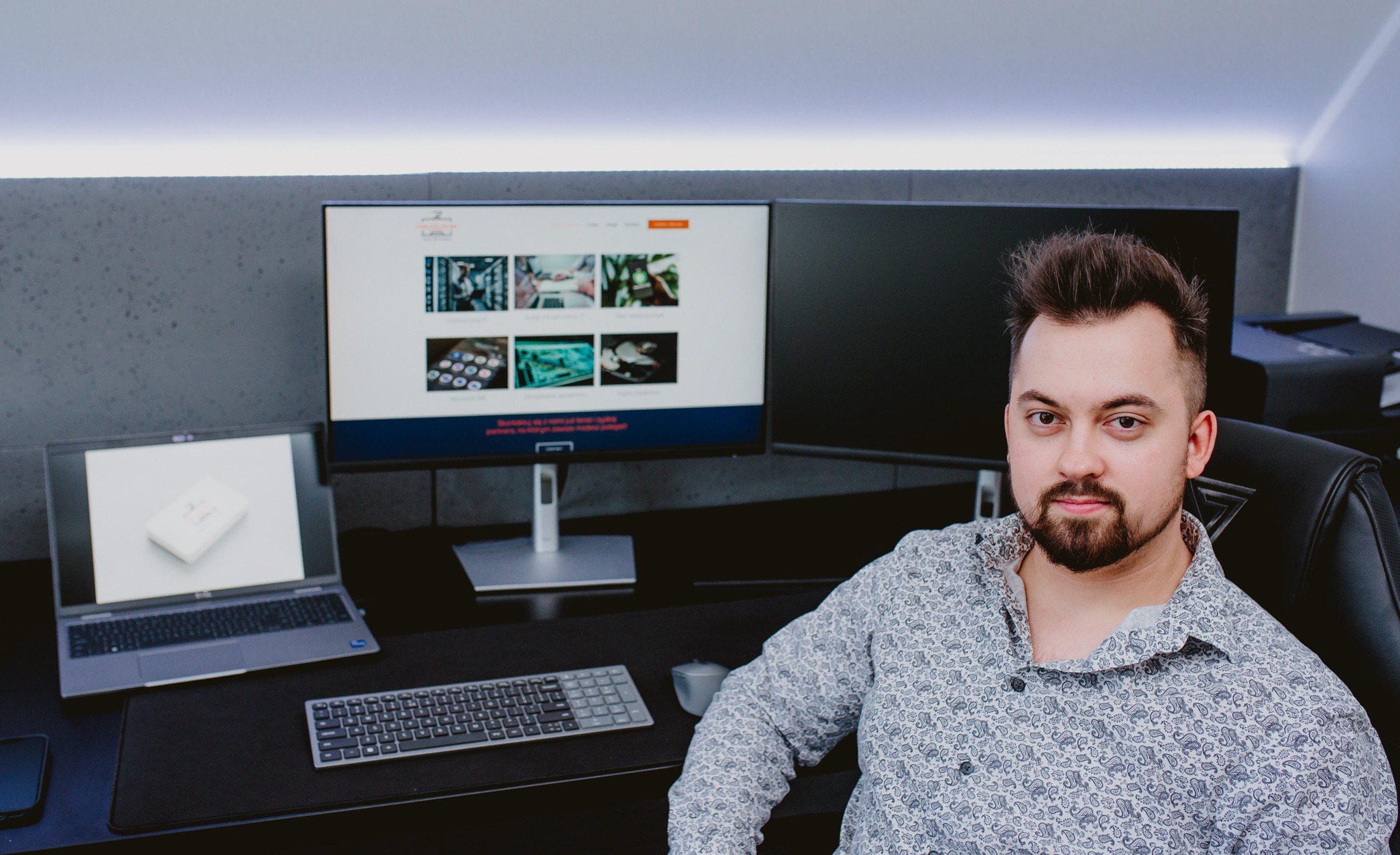 Mężczyzna siedzący przy biurku z dwoma monitorami, na jednym z nich widoczna strona internetowa firmy IT, obok laptop z logiem firmy na ekranie, klawiatura i myszka.