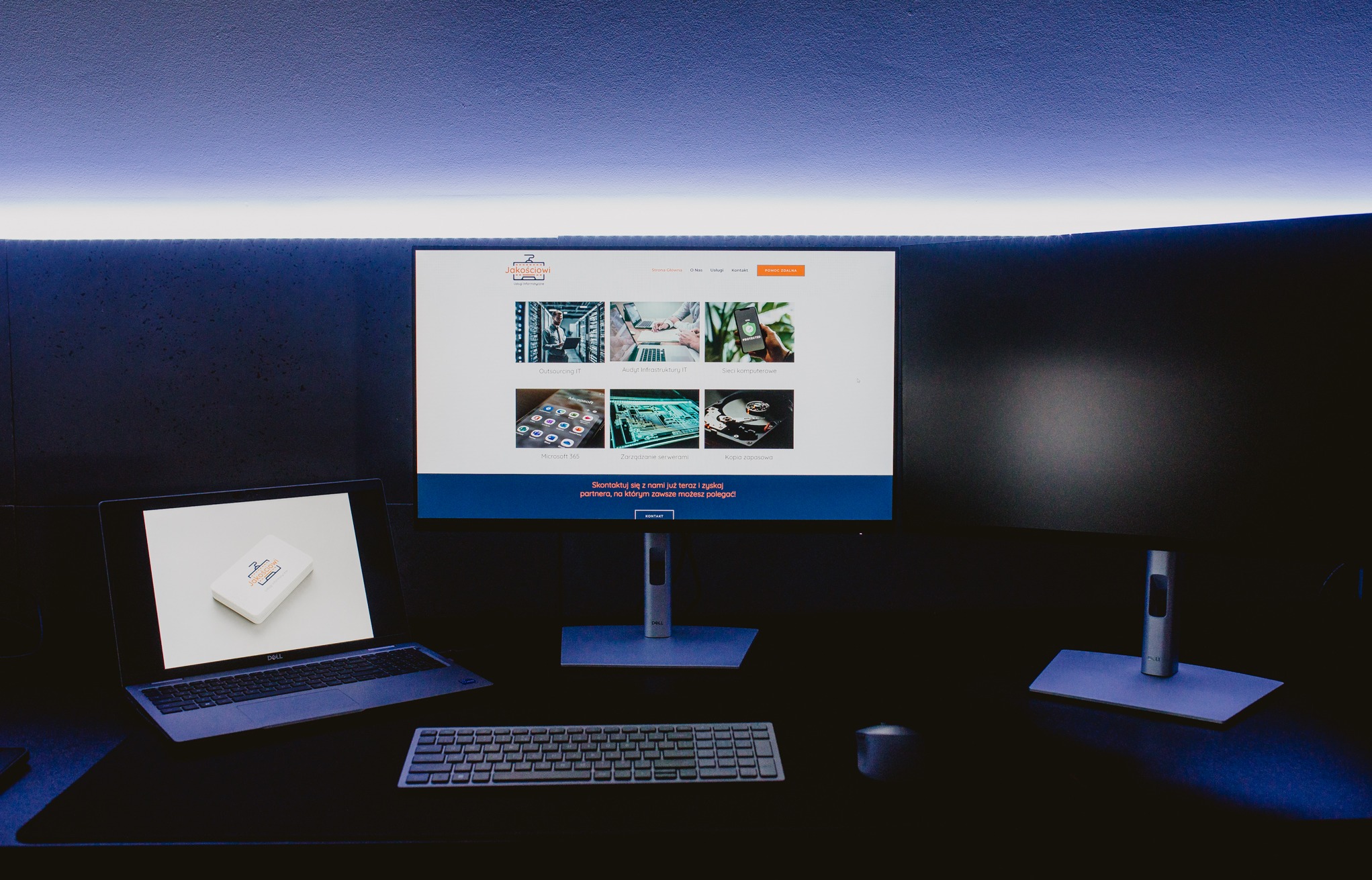 Biurko z laptopem Dell z wizytówką firmy IT na klapie, monitor wyświetla stronę internetową firmy IT z sekcjami Outsourcing IT, Audyt Infrastruktury IT i Sieci komputerowe, obok klawiatura i kubek.