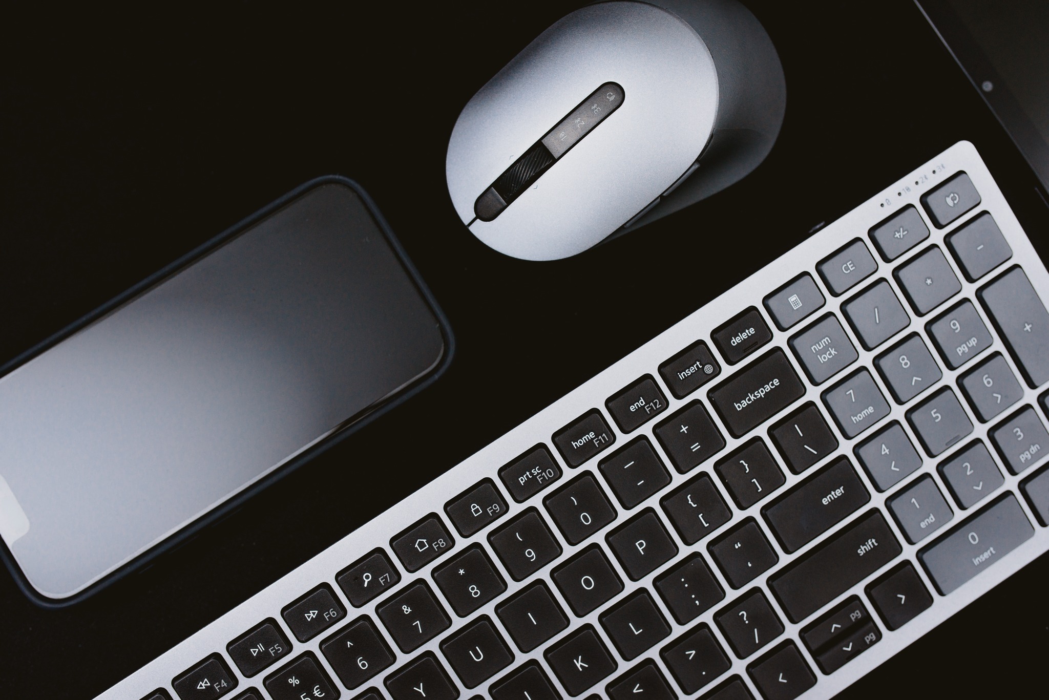 Minimalistyczne ujęcie z góry: srebrna klawiatura, mysz i smartfon na czarnym, błyszczącym biurku, tworzące nowoczesne stanowisko pracy.