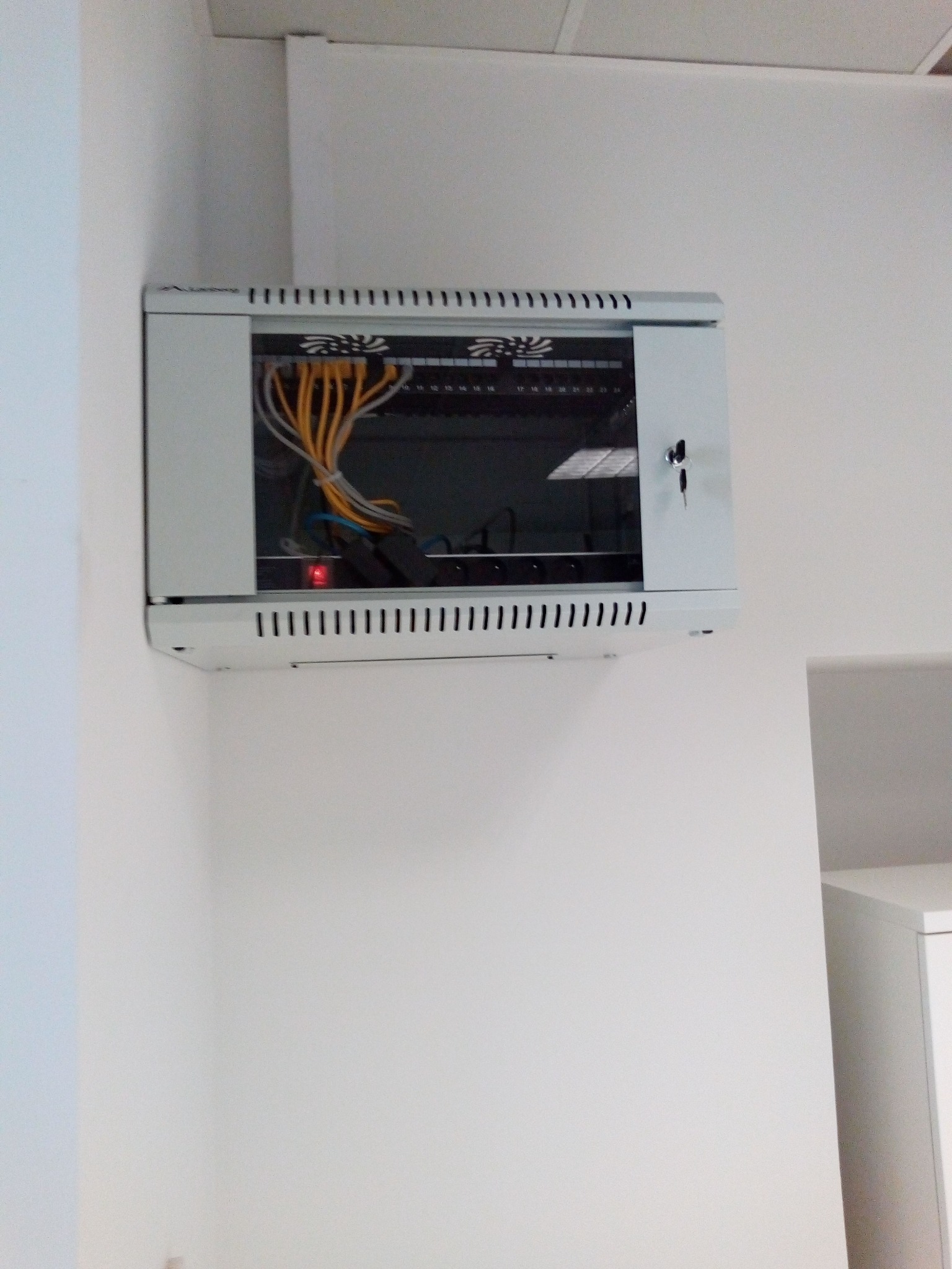 Zamknięta szafa rack wisząca na białej ścianie, widoczne kable sieciowe, na drzwiczkach szafy zamontowany zamek z kluczykiem.