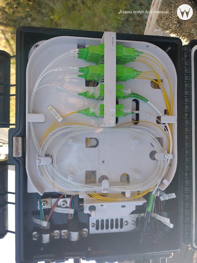 Otwarta skrzynka telekomunikacyjna z widocznymi światłowodami, złączami abonenckimi i OLT, oraz przewodami elektrycznymi w dolnej części, zamontowana na zewnątrz.