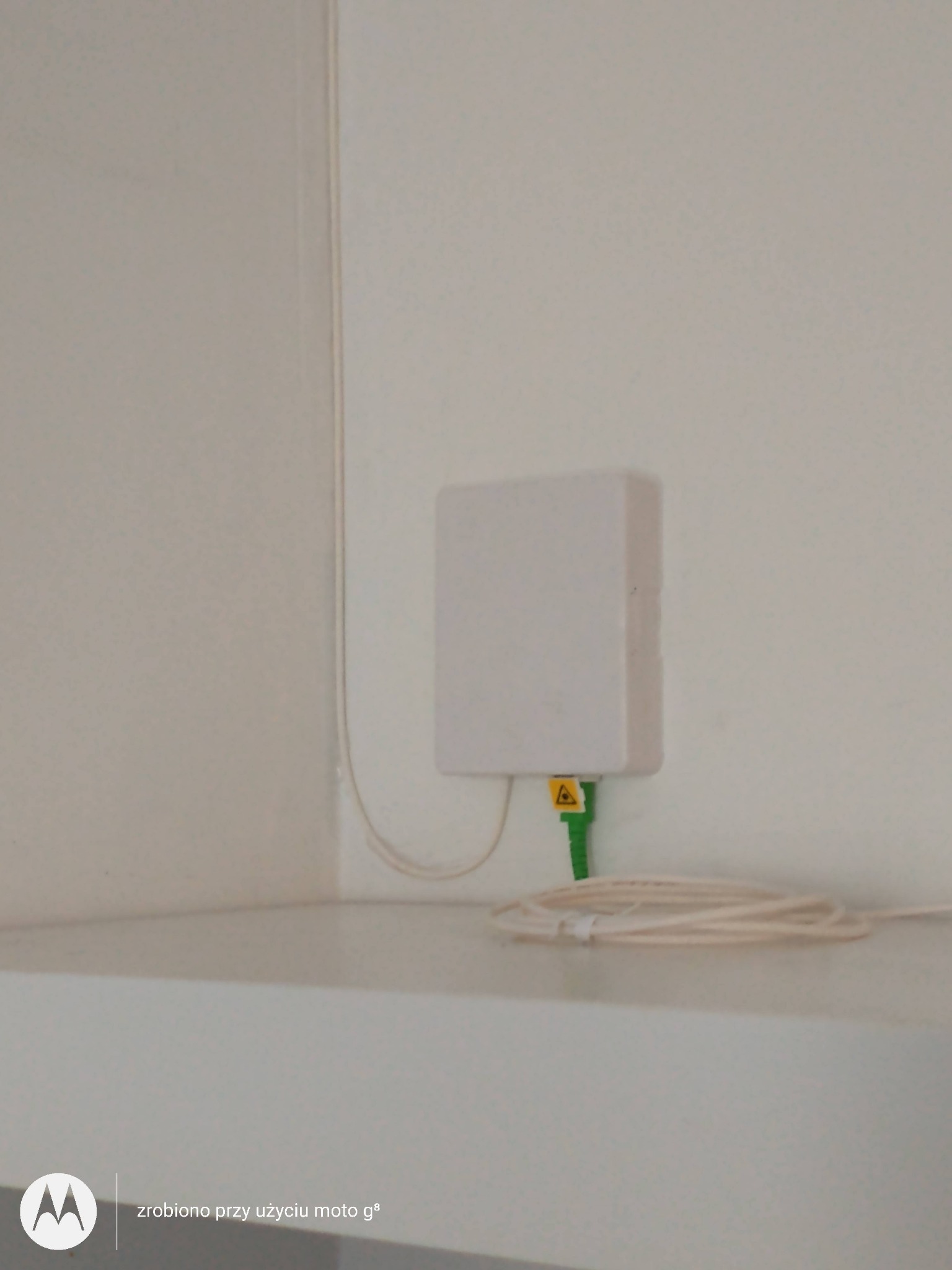 Biała skrzynka z kablem światłowodowym podłączona do ściany, widoczny żółty symbol lasera na złączu.