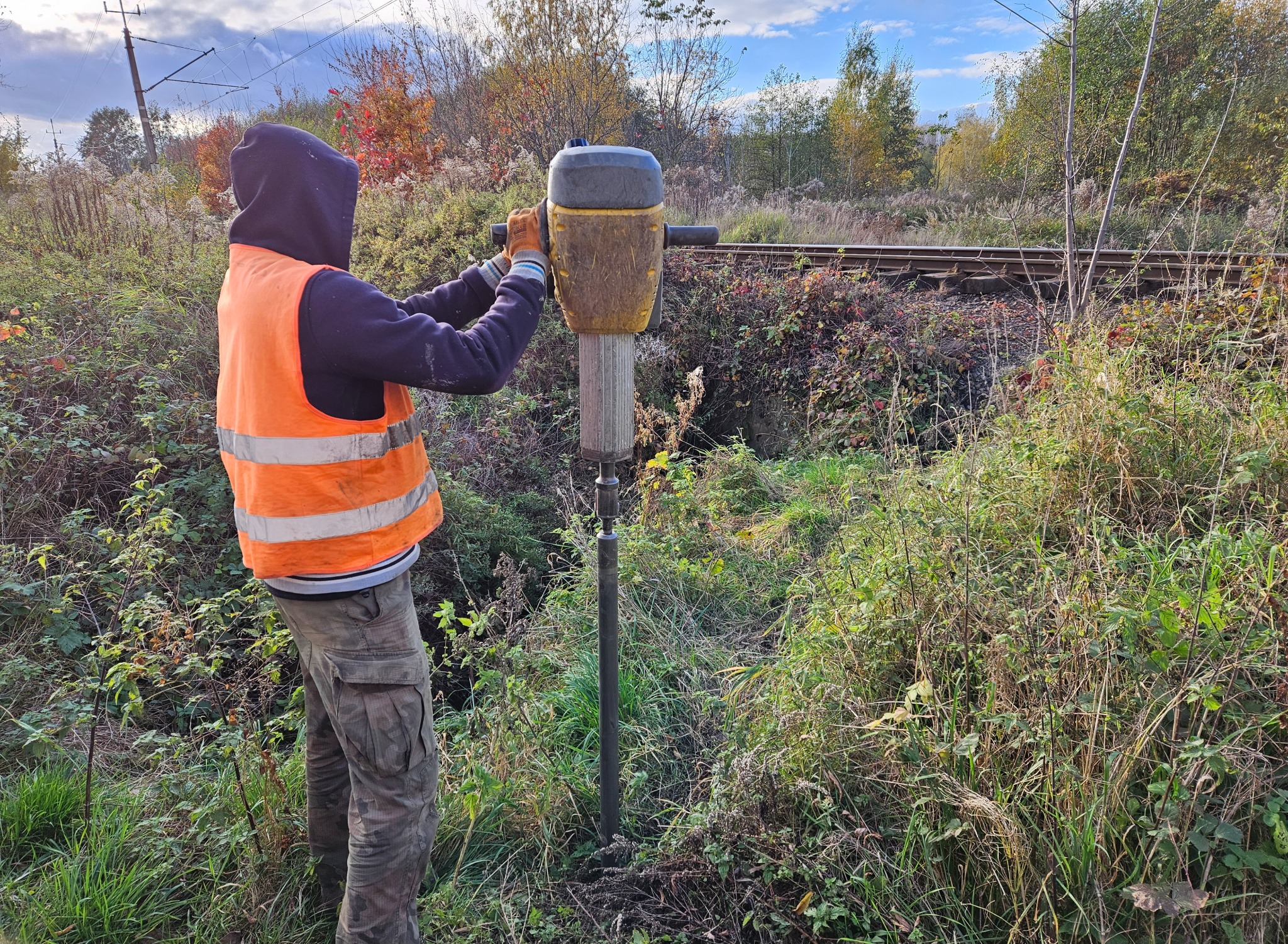 Pracownik w kamizelce odblaskowej wbija palik w ziemię przy torach kolejowych za pomocą wbijarki spalinowej w jesiennym krajobrazie.
