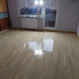 Świeżo polakierowany parkiet w pustym pokoju, zabezpieczone taśmą malarską krawędzie, widoczne odbicie lampy oświetleniowej na mokrej powierzchni.