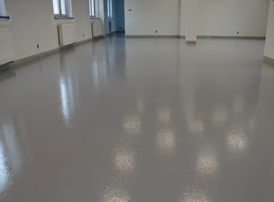 Gładka, szara posadzka w jasnym, przestronnym pomieszczeniu z białymi ścianami i grzejnikami. Powierzchnia podłogi odbija światło, tworząc subtelne refleksy. Przy ścianach widoczne zabezpieczenia...