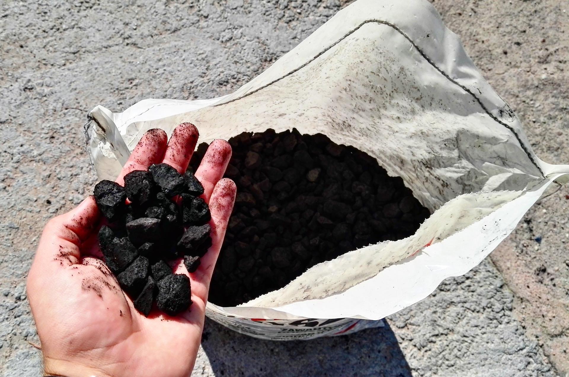 Dłoń trzyma bryłki ekogroszku na tle otwartego worka z ekogroszkiem na betonowej powierzchni.