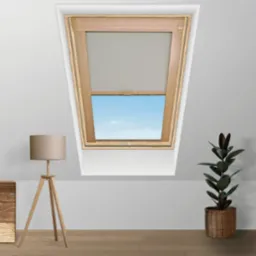 Okno dachowe z szarą roletą, drewniana rama, widok na błękitne niebo, obok lampa na trójnogu i roślina doniczkowa.