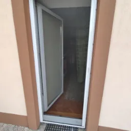 Otwarta moskitiera ramkowa w białej ramie, zamontowana w drzwiach wejściowych do domu z widokiem na wnętrze.