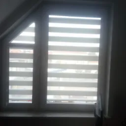 Okno na poddaszu z zamontowanymi roletami poziomymi z naprzemiennymi pasami transparentnymi i zaciemniającymi, widok z wnętrza pokoju.