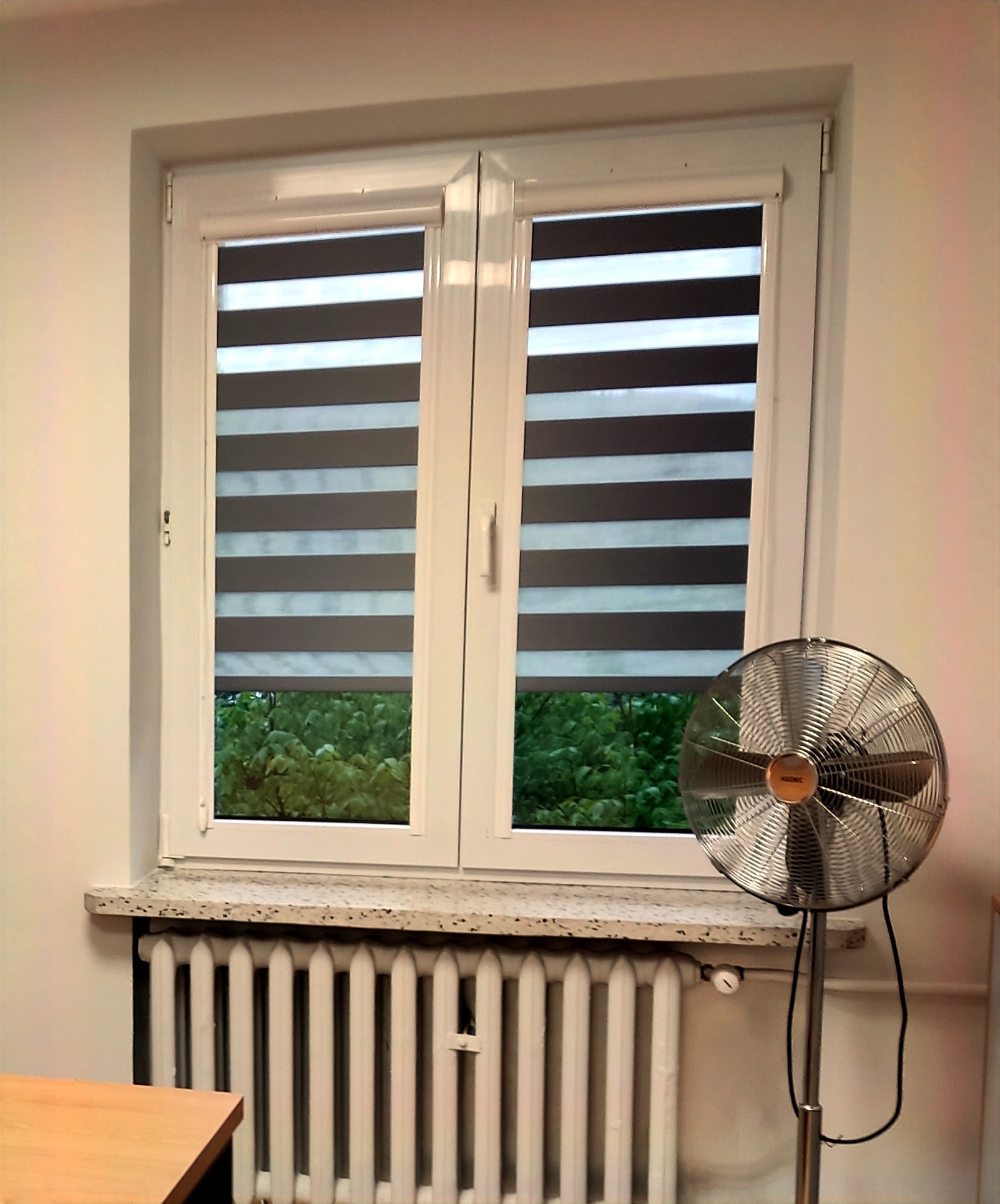Okno z podwójnymi skrzydłami, wyposażone w poziome rolety materiałowe w biało-szare pasy, za oknem widoczna zieleń, na pierwszym planie metalowy wentylator stojący.
