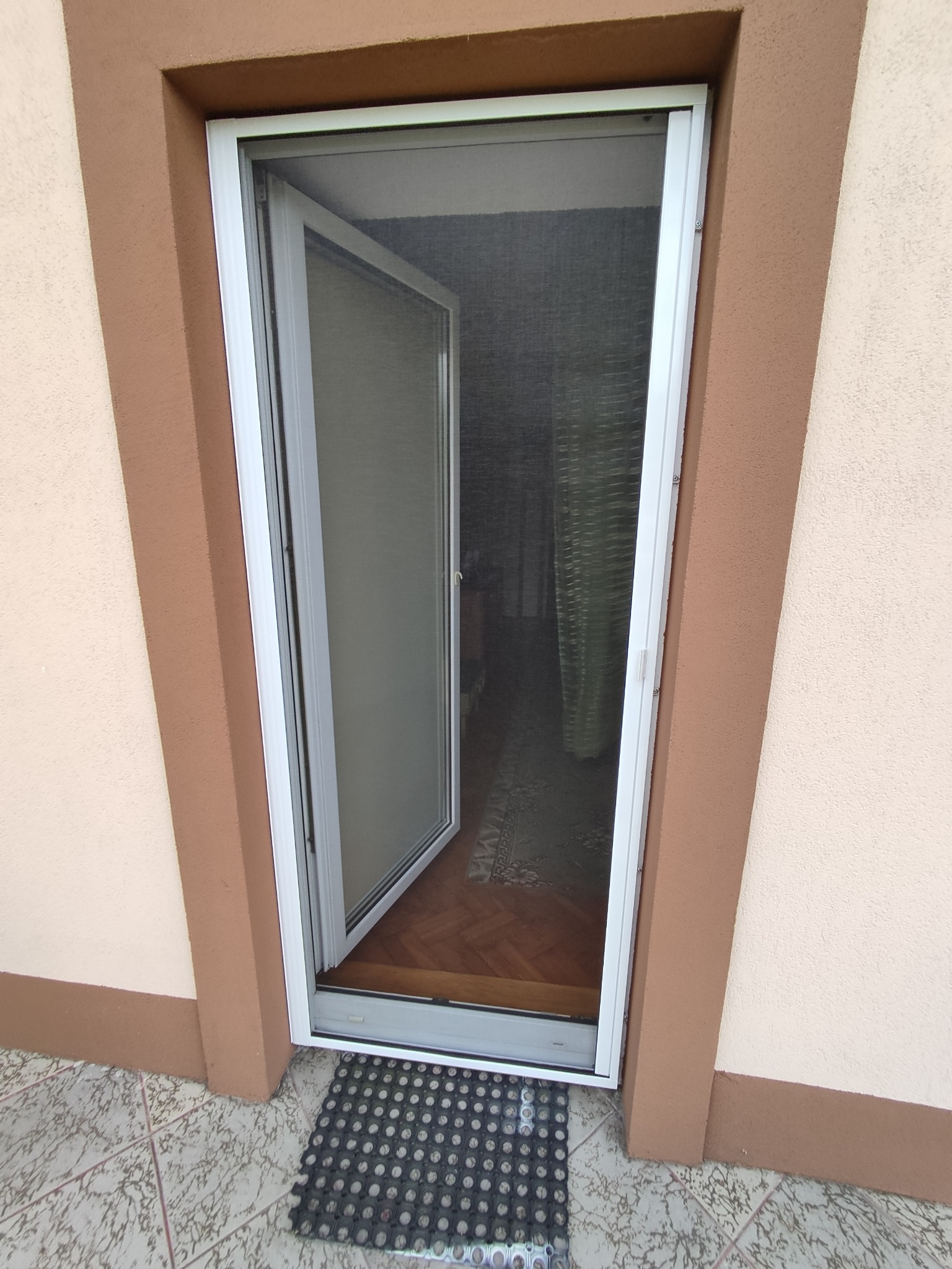 Otwarta moskitiera ramkowa w białej ramie, zamontowana w drzwiach wejściowych do domu z widokiem na wnętrze.