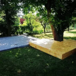 Nowo wybudowany, surowy drewniany taras w kształcie litery L, sąsiadujący z przygotowanym podłożem z szarego żwiru, w zielonym ogrodzie z drzewami.