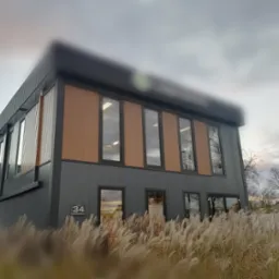 Nowoczesny budynek z szarą elewacją i drewnianymi panelami, widoczny z perspektywy niskich traw ozdobnych na pierwszym planie. Na budynku widnieje numer '34 Nowosielec'.