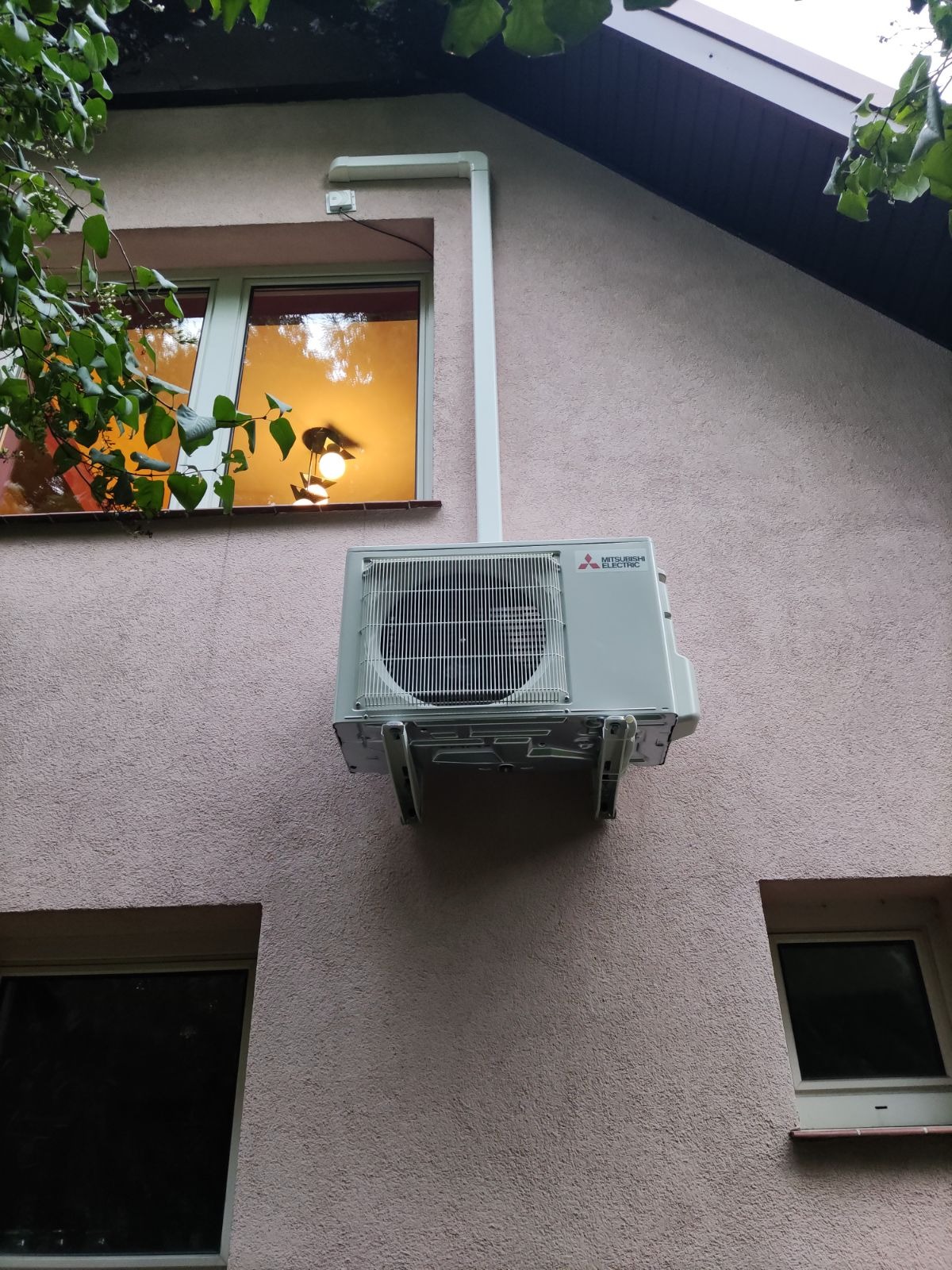 Zewnętrzna jednostka klimatyzacji Mitsubishi Electric zamontowana na różowej elewacji domu, z widocznymi oknami i fragmentami rynny.