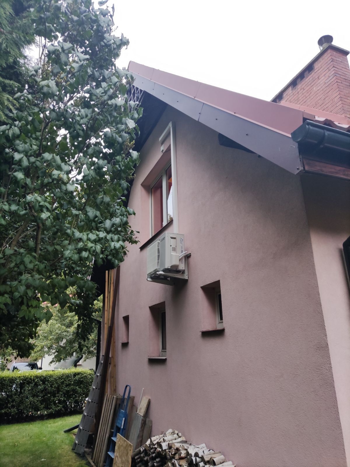 Zewnętrzna jednostka klimatyzacji zamontowana na elewacji domu w Duczkach, widoczna z boku, obok okna i sterty drewna.