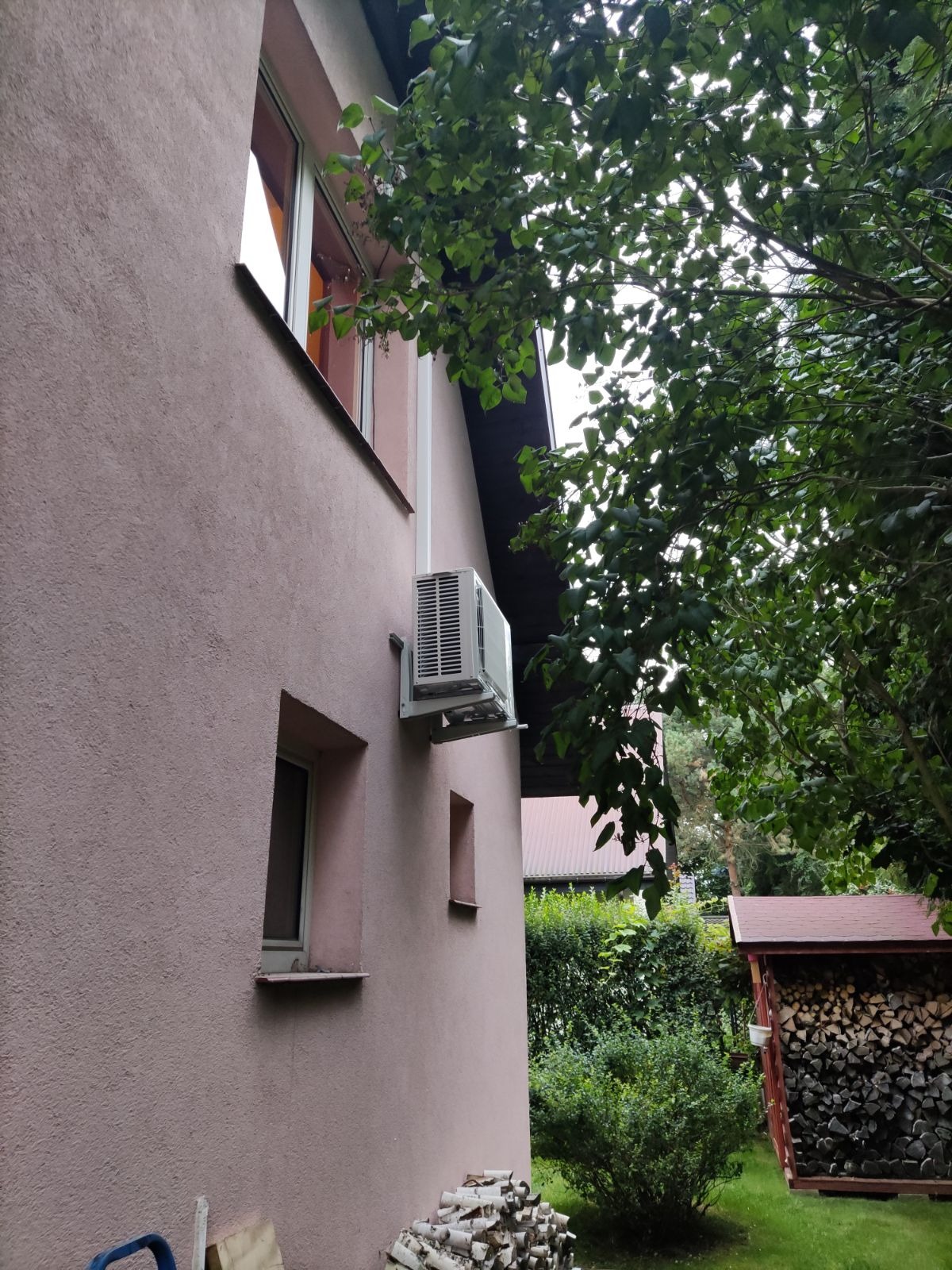 Zewnętrzna jednostka klimatyzacji zamontowana na ścianie domu o różowej elewacji, widok z boku, z widocznymi oknami, stosem drewna i zielenią w tle.