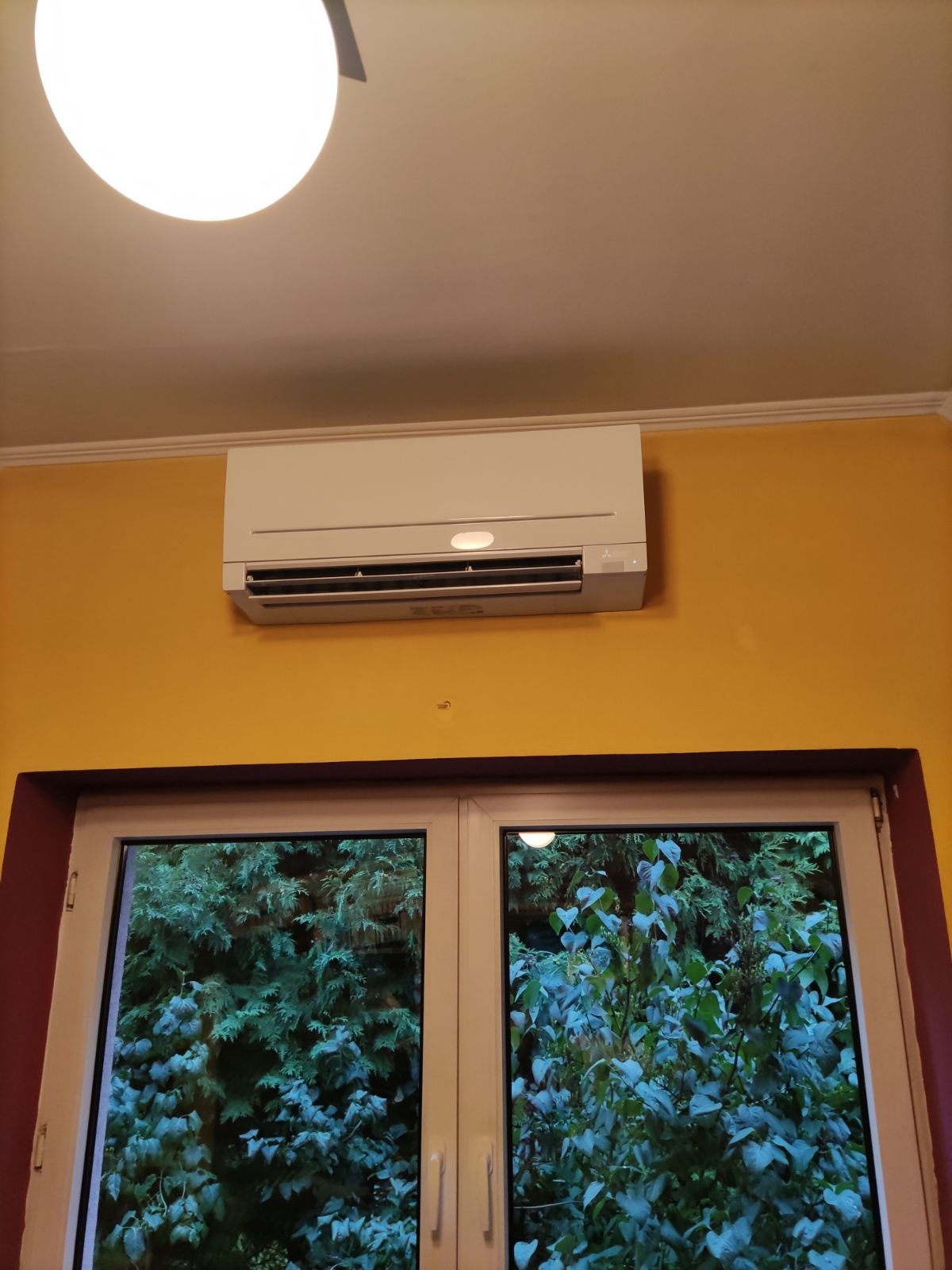 Biała jednostka klimatyzacji Mitsubishi Electric zamontowana na żółtej ścianie nad oknem z widokiem na zielone drzewa.