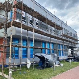 Marlena Grablowska - Budynek w trakcie ocieplania elewacji, osłonięte okna folią, widoczne rusztowania i materiały budowlane na trawniku, pochmurne niebo.