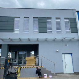 Marlena Grablowska - Mężczyzna maluje elewację budynku na szaro, widoczne puszki z farbą, podnośnik koszowy w tle, ulica Nowy Świat 40.