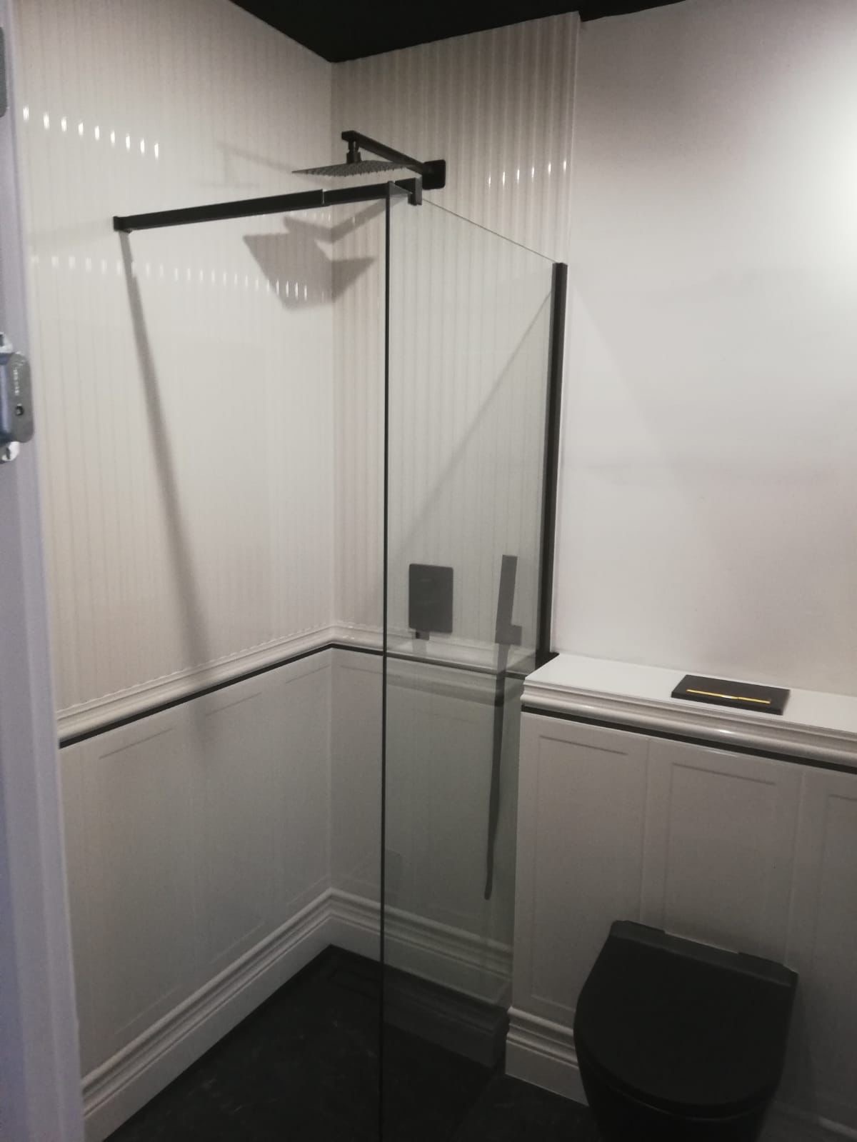 Nowoczesna łazienka z czarną armaturą: kabina prysznicowa typu walk-in z deszczownicą, białe panele ścienne z czarnym akcentem, czarna toaleta wisząca i czarny sufit.