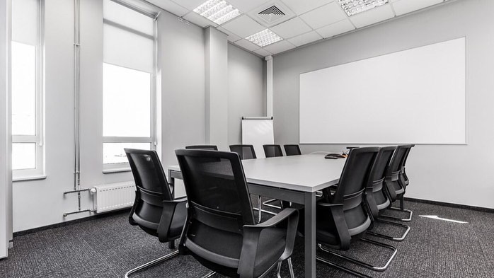 Nowoczesne biuro z szarą wykładziną, białymi ścianami, oknami z roletami, dużym stołem konferencyjnym z czarnymi krzesłami i tablicą suchościeralną.