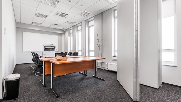 Nowoczesne biuro w Gdyni z szarą wykładziną dywanową, białymi ścianami i sufitem podwieszanym z rastrowym oświetleniem. Wnętrze wyposażone w drewniane biurka na metalowych nogach, czarne krzesła...
