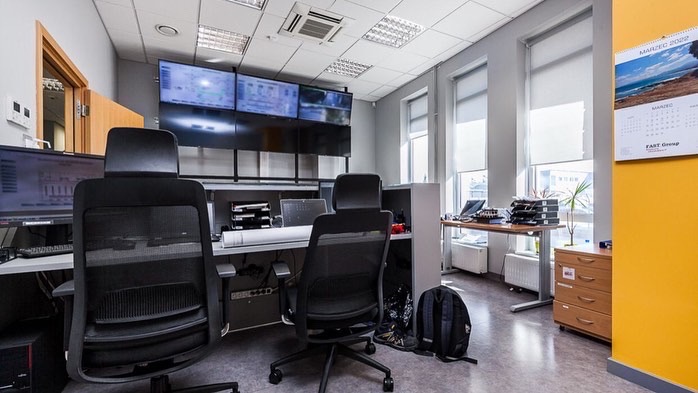 Jasne biuro z widocznymi stanowiskami pracy, monitorami prezentującymi schematy i planami architektonicznymi, kalendarzem na ścianie i elementami wyposażenia biurowego.