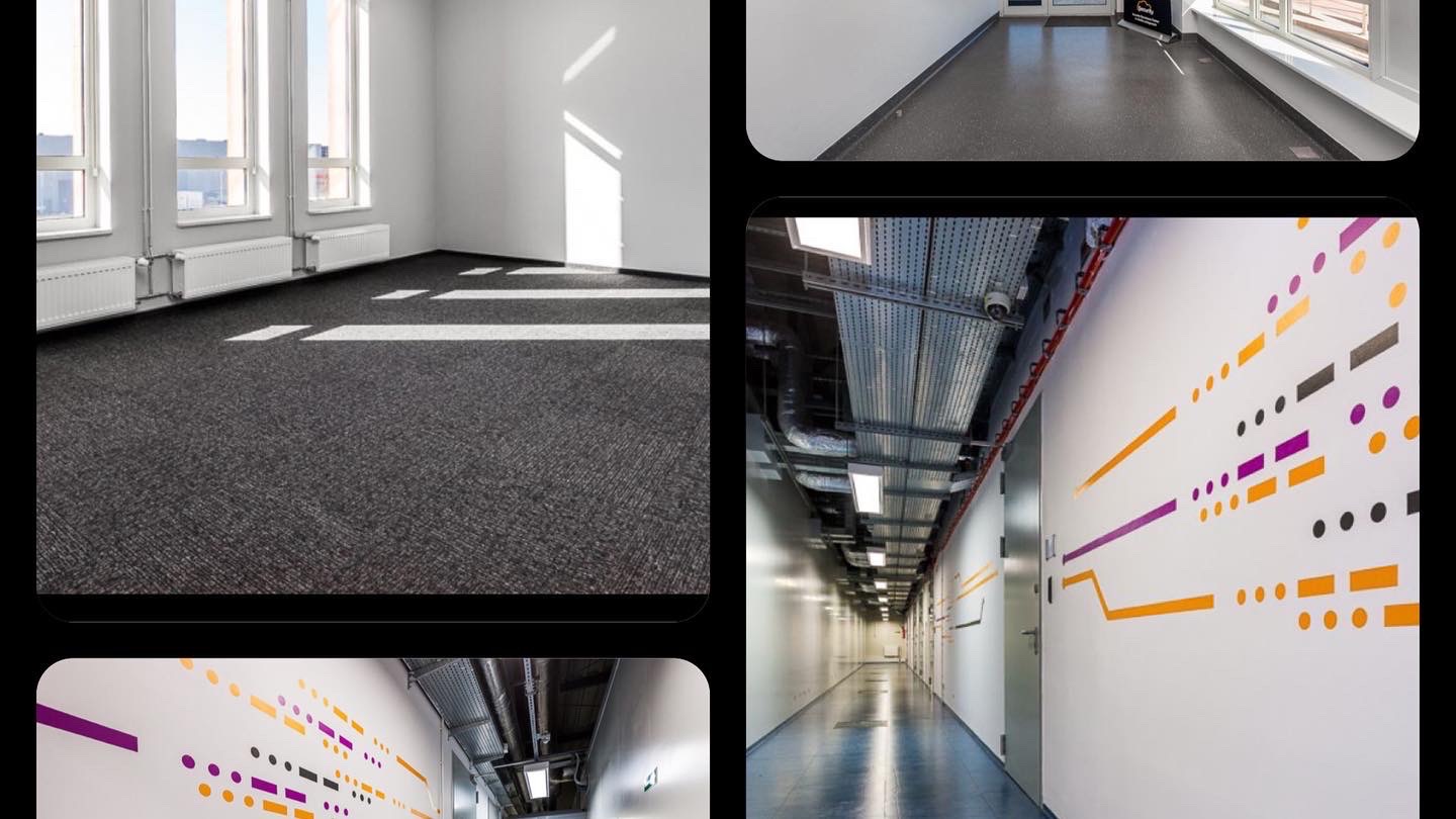 Wnętrze biurowe z nową wykładziną dywanową w odcieniach szarości, białe ściany z dekoracyjnymi wzorami w kolorach pomarańczowym i fioletowym, oraz widoczne instalacje pod sufitem.