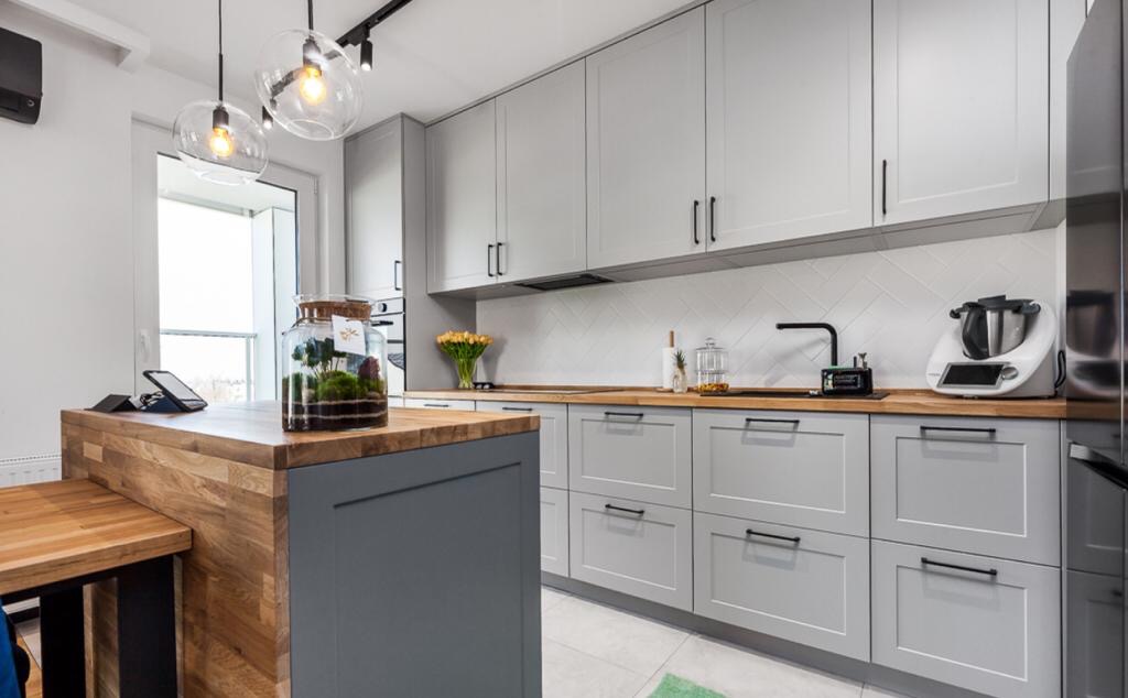 Jasna kuchnia w Gdyni z szarymi szafkami, drewnianym blatem i wyspą, oświetlona wiszącymi lampami, z dekoracyjnym terrarium na blacie.