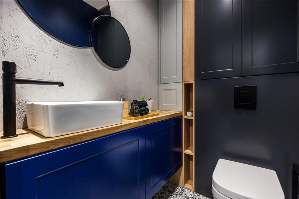 Nowoczesna łazienka z niebieskimi szafkami, drewnianym blatem, białą umywalką i czarną armaturą na tle szarej ściany z okrągłymi lustrami.