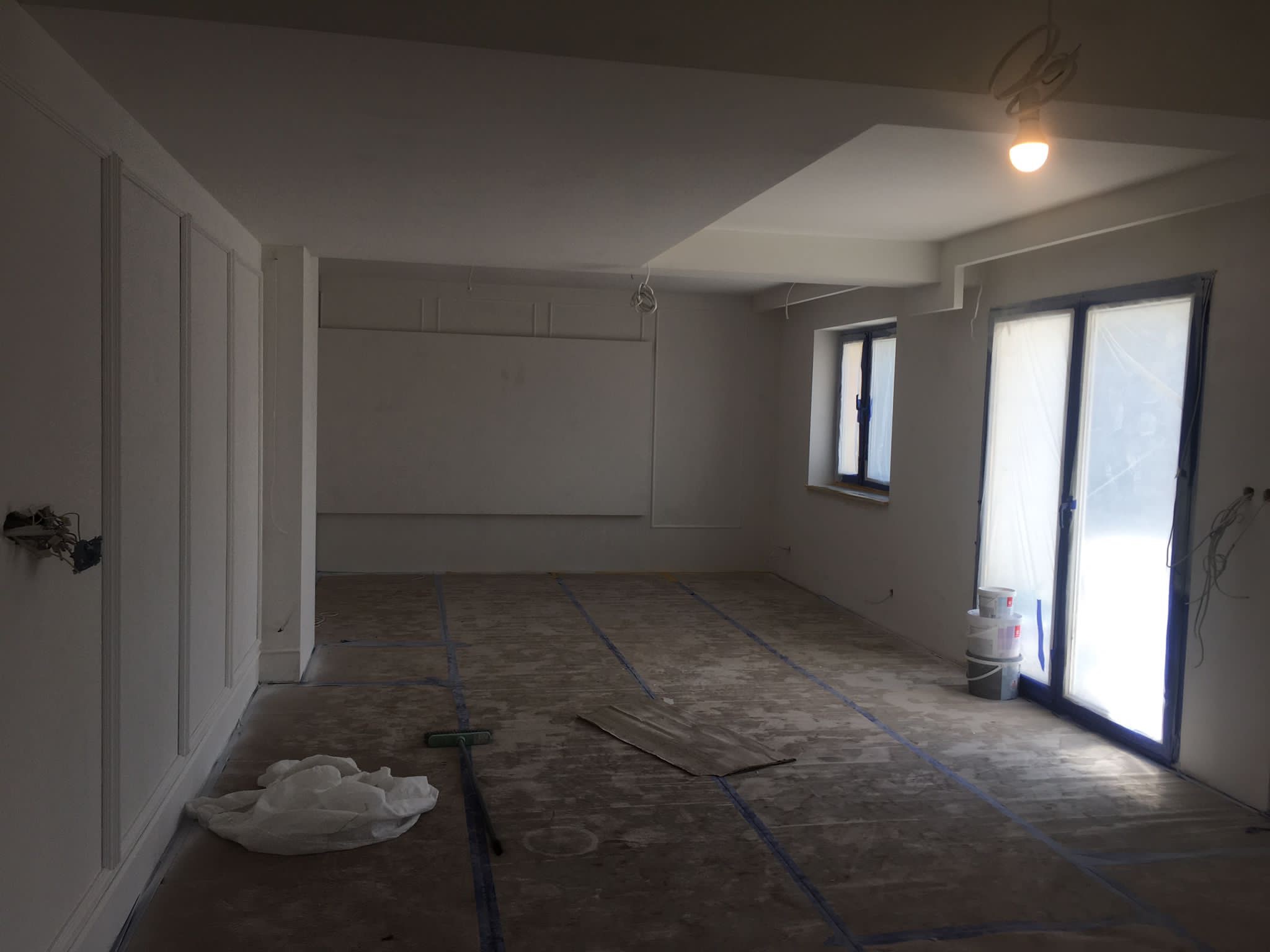 Wnętrze w trakcie remontu: białe ściany z dekoracyjnymi panelami, zabezpieczone okna folią, przygotowane podłoże pod panele, widoczne puszki z farbą i narzędzia.