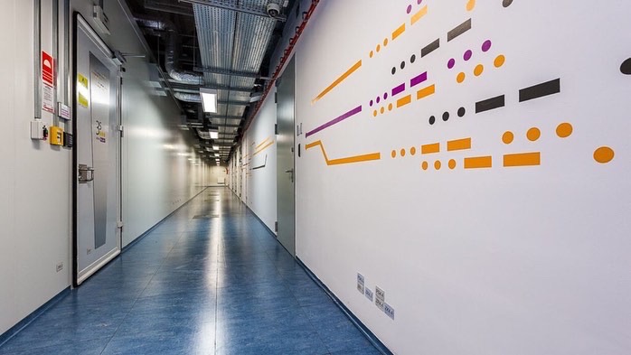 Długi korytarz w budynku przemysłowym z niebieską podłogą, białymi ścianami ozdobionymi abstrakcyjnym wzorem z kropek i linii w kolorach pomarańczowym, fioletowym i czarnym, widoczne instalacje...