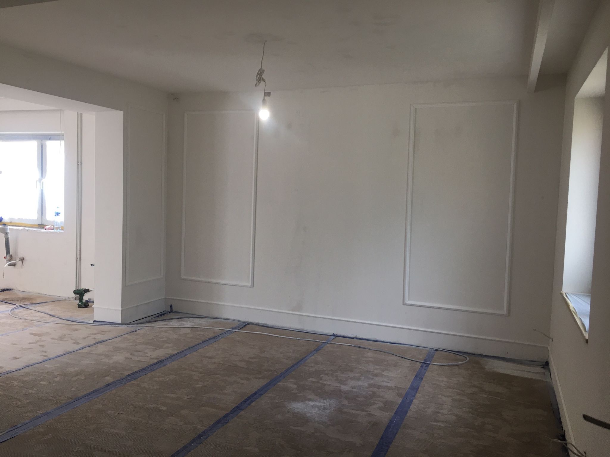 Wnętrze pokoju w trakcie prac wykończeniowych, widoczne listwy dekoracyjne na ścianach, zabezpieczona podłoga i wisząca żarówka.
