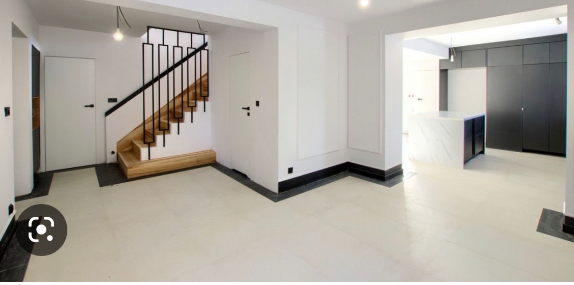 Jasne, przestronne wnętrze domu po remoncie z drewnianymi schodami z czarną balustradą, białymi ścianami z czarnymi listwami przypodłogowymi i podłogą w odcieniu kremowym, widok na kuchnię z wyspą...