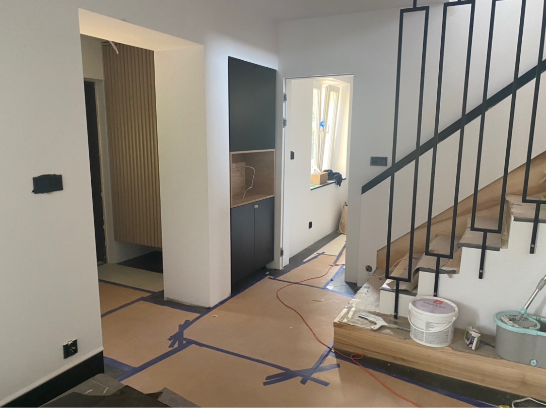 Wnętrze w trakcie prac wykończeniowych: zabezpieczona podłoga kartonem i taśmą, widoczne schody z czarną balustradą, zabudowa meblowa w kolorze czarnym i naturalnego drewna, wiadro z farbą oraz mop.