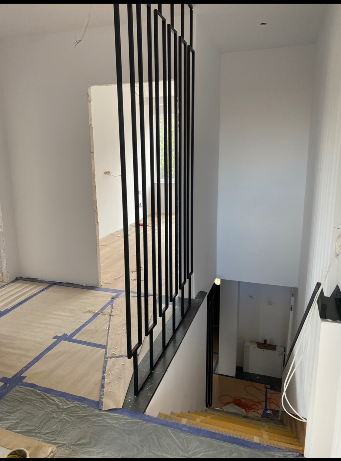 Wnętrze w trakcie prac wykończeniowych: widok na schody z czarną, metalową balustradą z pionowych prętów, zabezpieczona podłoga kartonem i folią malarską, surowe ściany przygotowane do malowania.