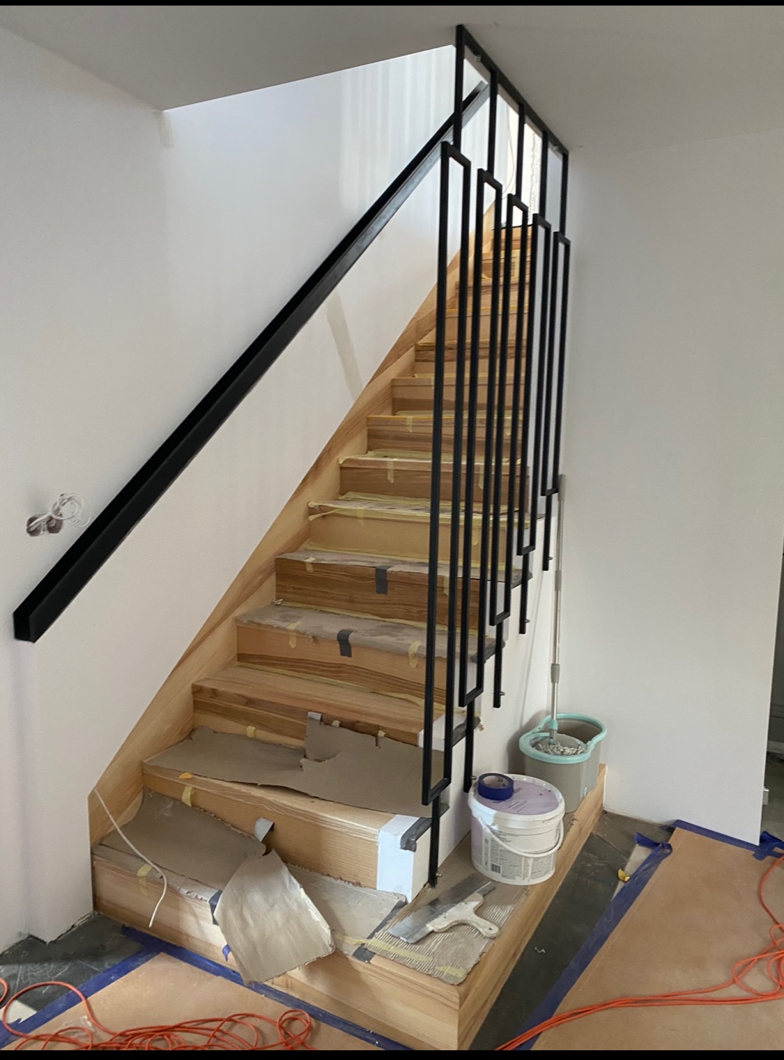 Montaż drewnianych schodów z nowoczesną, czarną balustradą w trakcie prac wykończeniowych, zabezpieczone folią i taśmą malarską, widoczne narzędzia i wiadro z farbą.