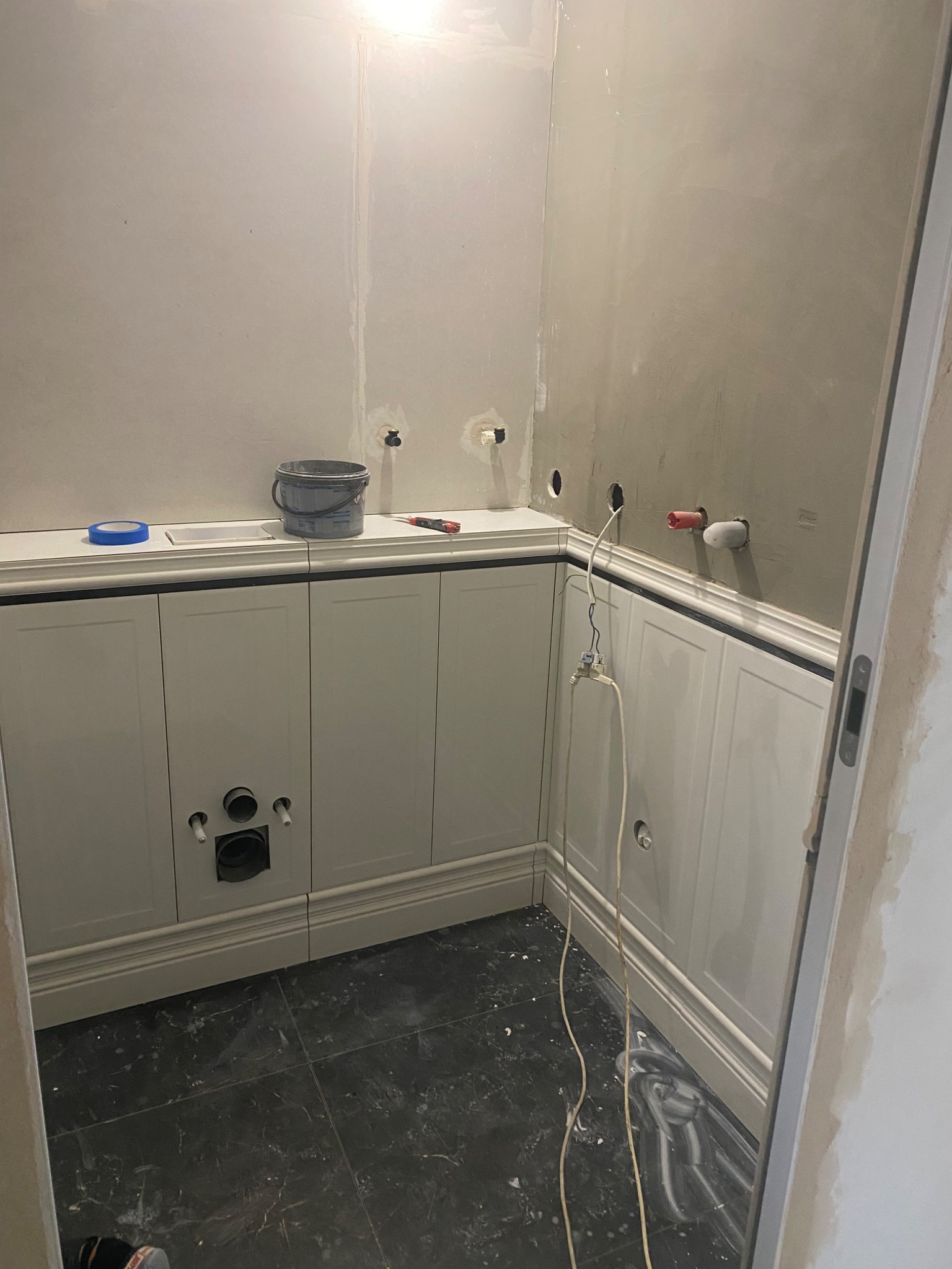 Łazienka w trakcie wykończenia: białe panele ścienne z czarnymi akcentami, czarna marmurowa podłoga, przygotowane punkty instalacyjne, widoczne narzędzia i wiadro farby.