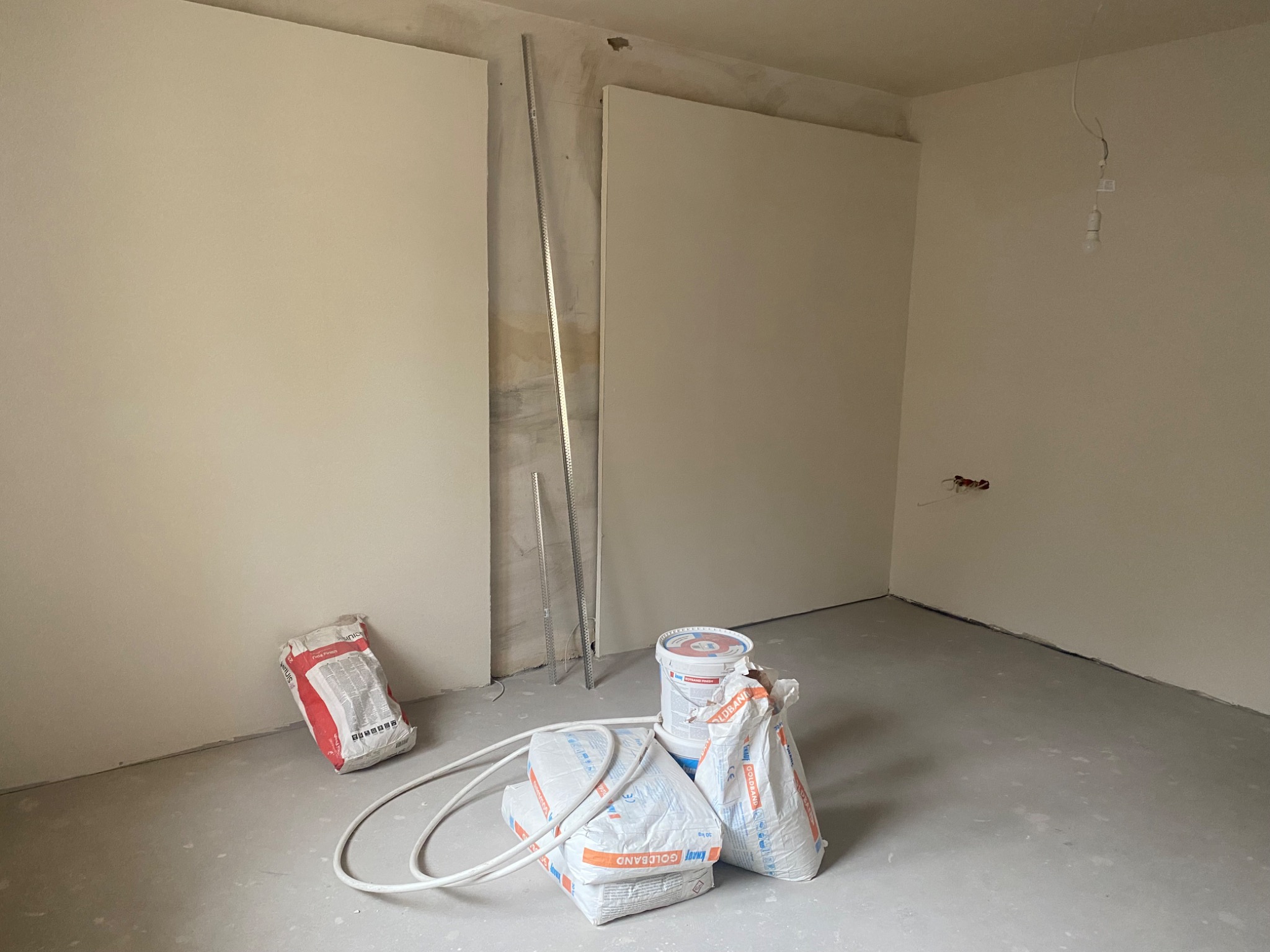 Wnętrze w trakcie prac wykończeniowych: widoczne płyty kartonowo-gipsowe na ścianach, profile, worki z gładzią gipsową Knauf Goldband oraz wiadro z masą szpachlową, surowe oświetlenie żarówką...