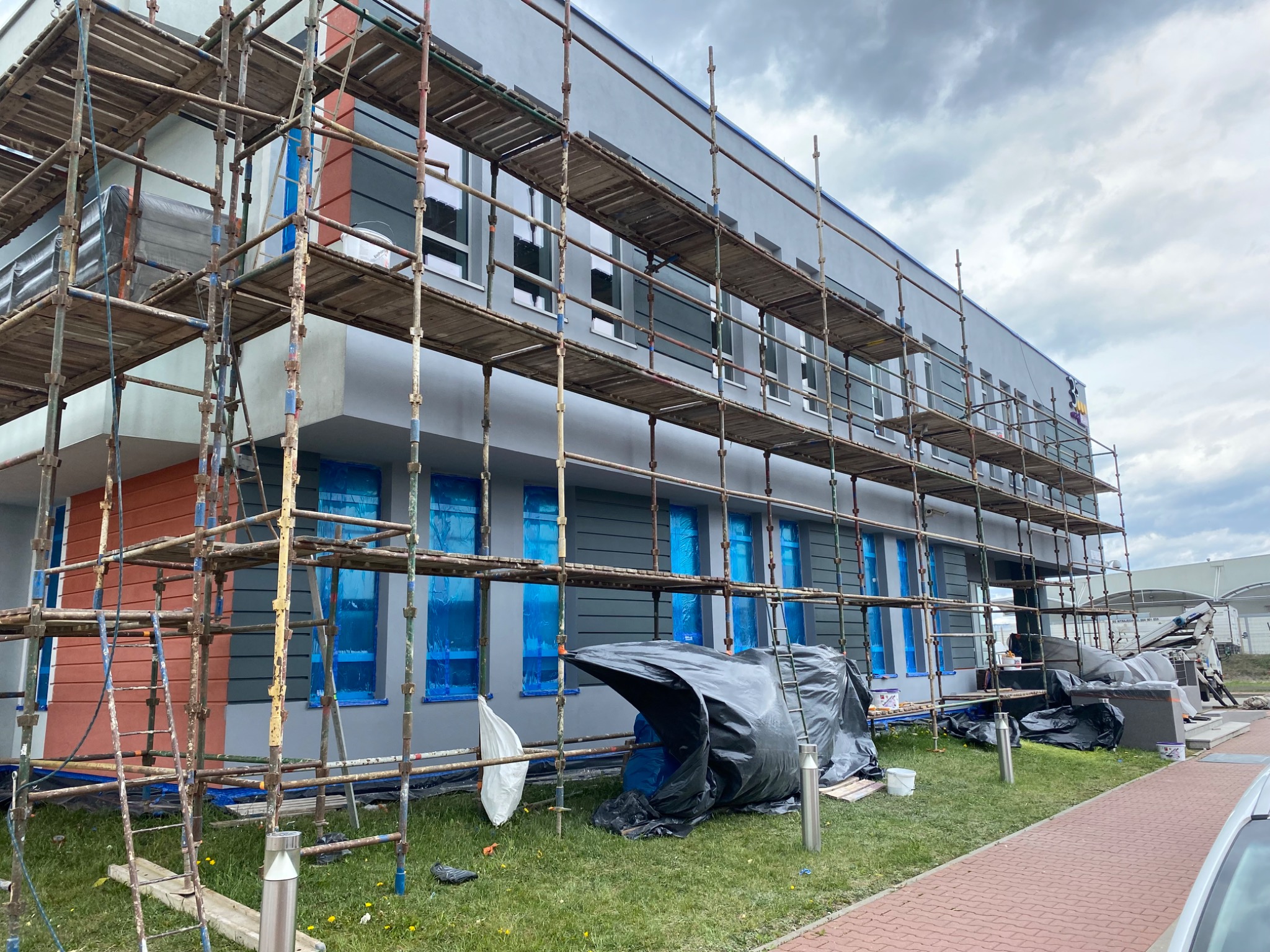 Budynek w trakcie ocieplania elewacji, osłonięte okna folią, widoczne rusztowania i materiały budowlane na trawniku, pochmurne niebo.