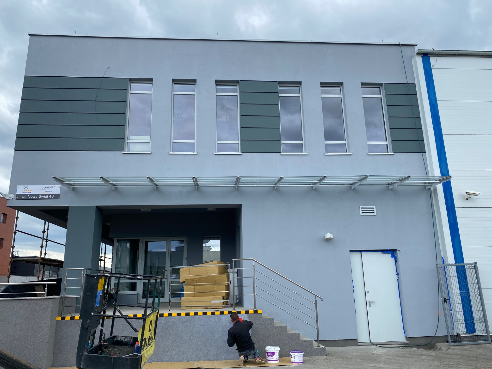 Mężczyzna maluje elewację budynku na szaro, widoczne puszki z farbą, podnośnik koszowy w tle, ulica Nowy Świat 40.