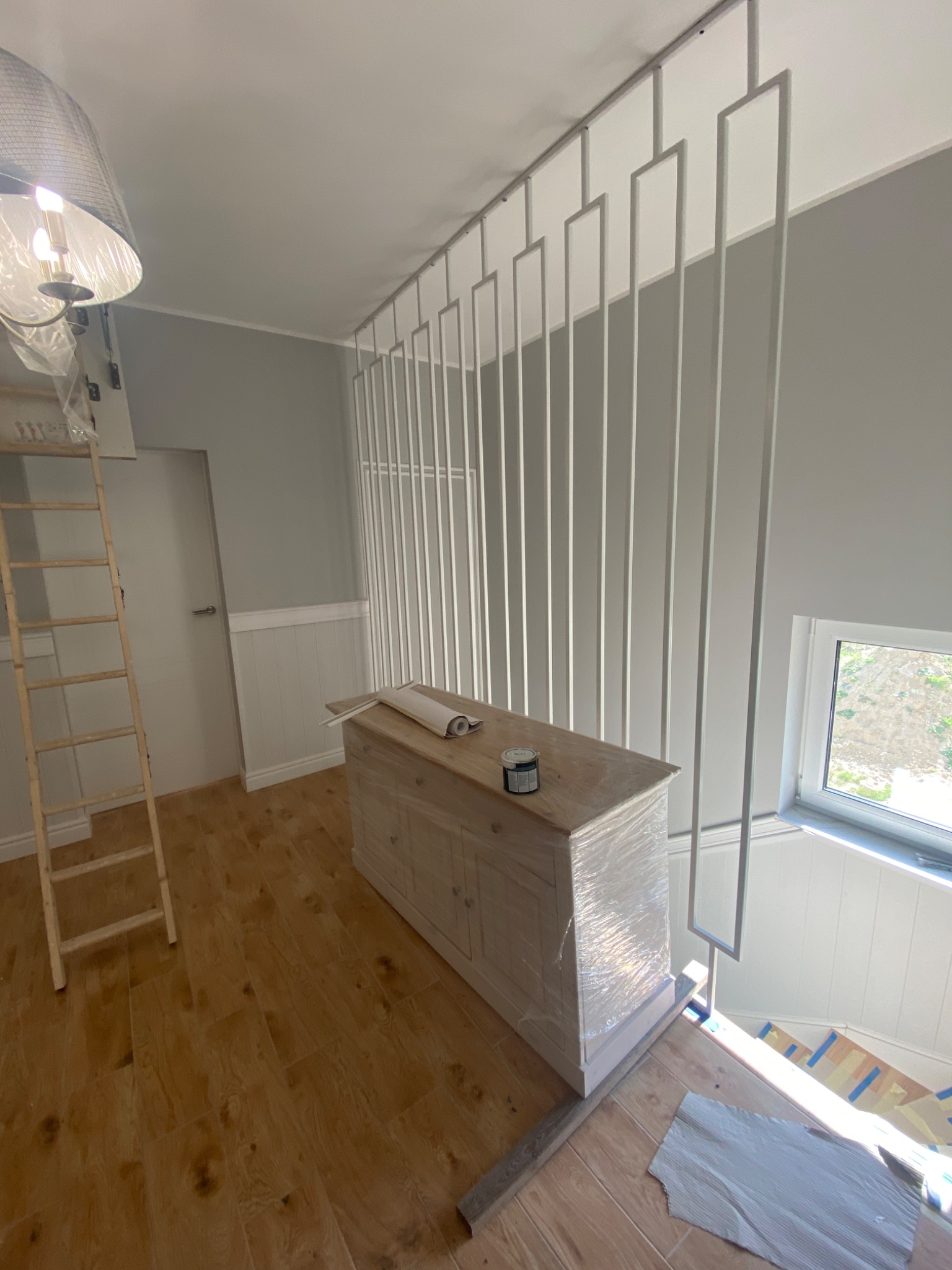 Wnętrze w trakcie wykańczania: drewniana komoda owinięta folią, puszka farby, częściowo położone panele na schodach, dekoracyjna, biała konstrukcja sufitowa z pionowych elementów, drabina oparta...