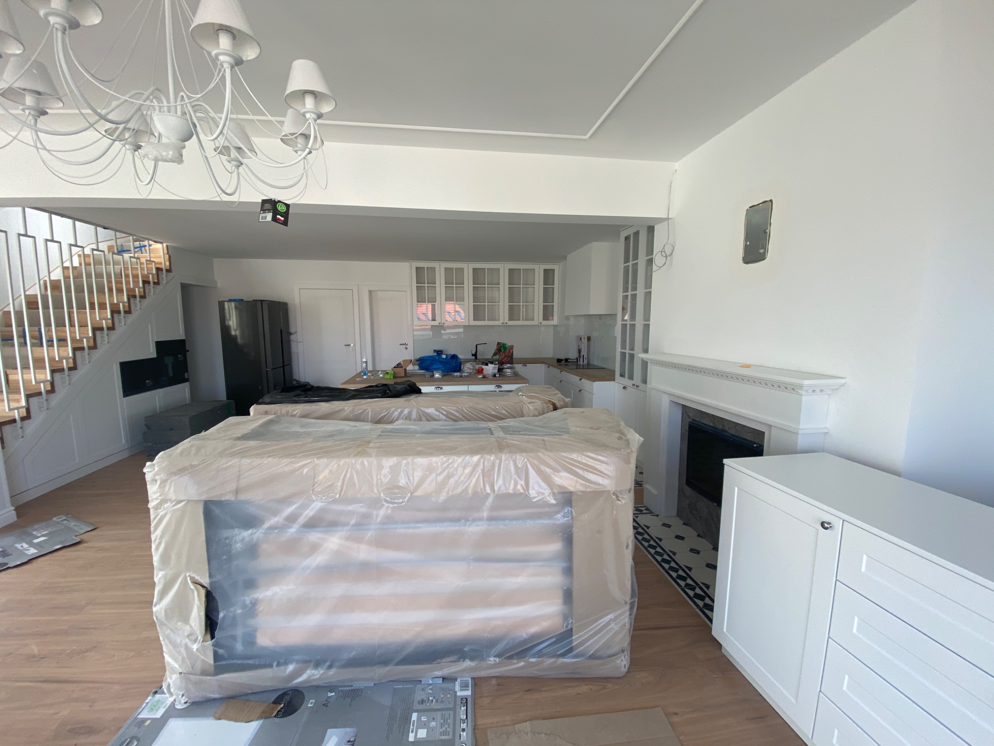 Wnętrze w trakcie wykończenia: zabezpieczone folią meble na podłodze z paneli, białe ściany z przygotowanym miejscem na obraz, kominek, schody z białymi balustradami i designerski żyrandol.
