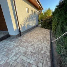 Ułożona kostka brukowa w odcieniach beżu i brązu tworząca chodnik przy domu z jasną elewacją i brązową rynną, widoczne krzewy i drzewa w tle.