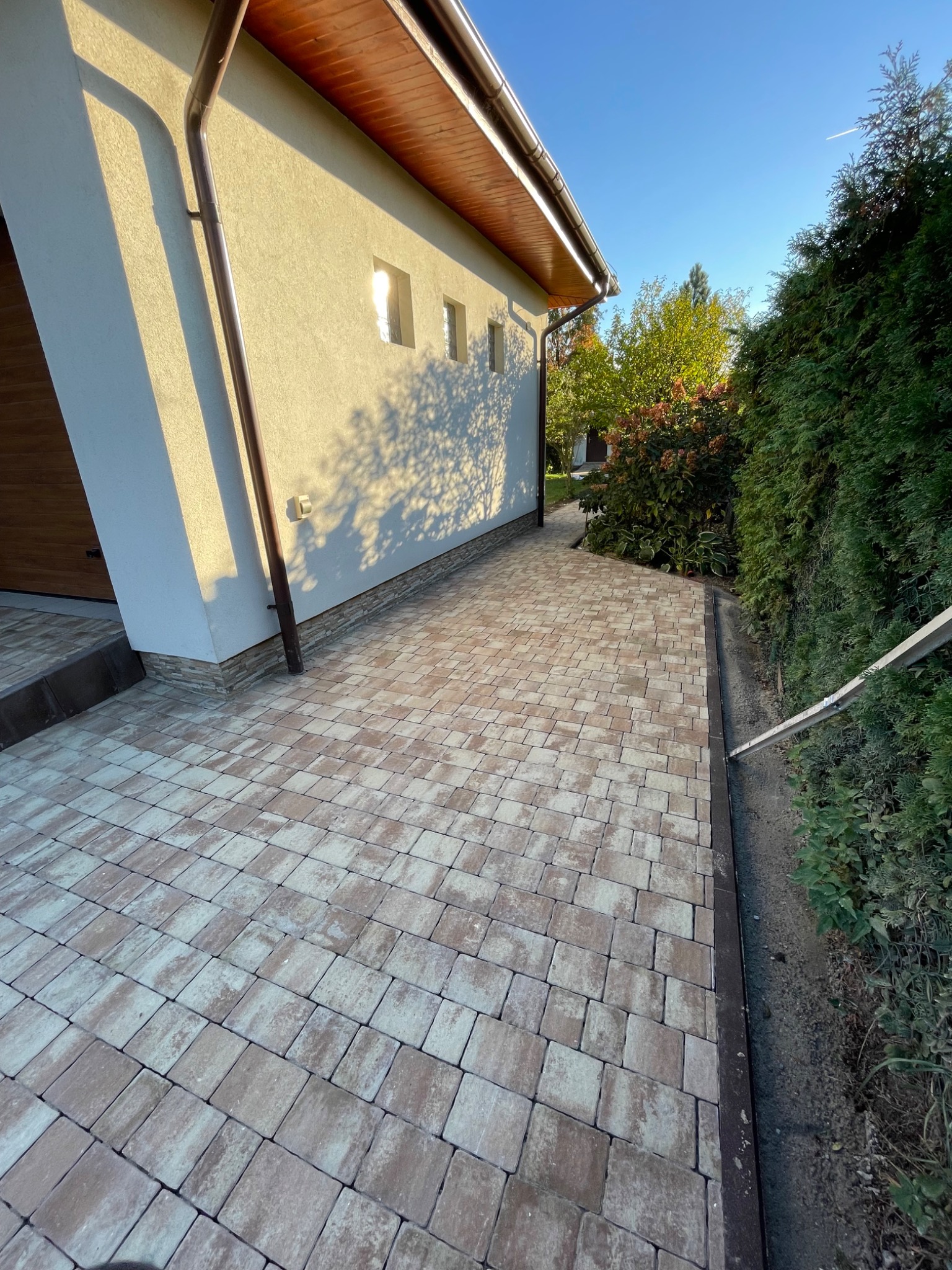 Ułożona kostka brukowa w odcieniach beżu i brązu tworząca chodnik przy domu z jasną elewacją i brązową rynną, widoczne krzewy i drzewa w tle.