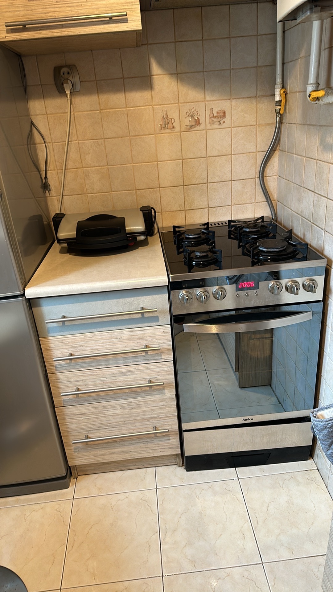 Fragment kuchni z płytą gazową, piekarnikiem Amica, szafką z szufladami i grillem elektrycznym na blacie, widoczne elementy instalacji gazowej i gniazdko elektryczne.