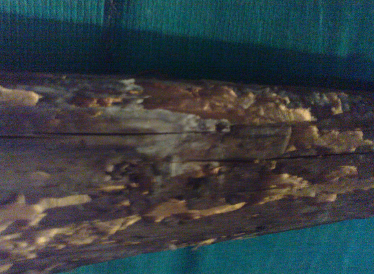 Silnie uszkodzona, spróchniała belka drewniana z widocznymi śladami gnicia i ubytkami, na tle turkusowej tkaniny.