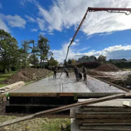 Wylewanie betonu na przygotowany fundament domu jednorodzinnego z użyciem pompy do betonu; widoczni pracownicy w trakcie rozprowadzania betonu, zbrojenie w fundamencie, stos desek szalunkowych...