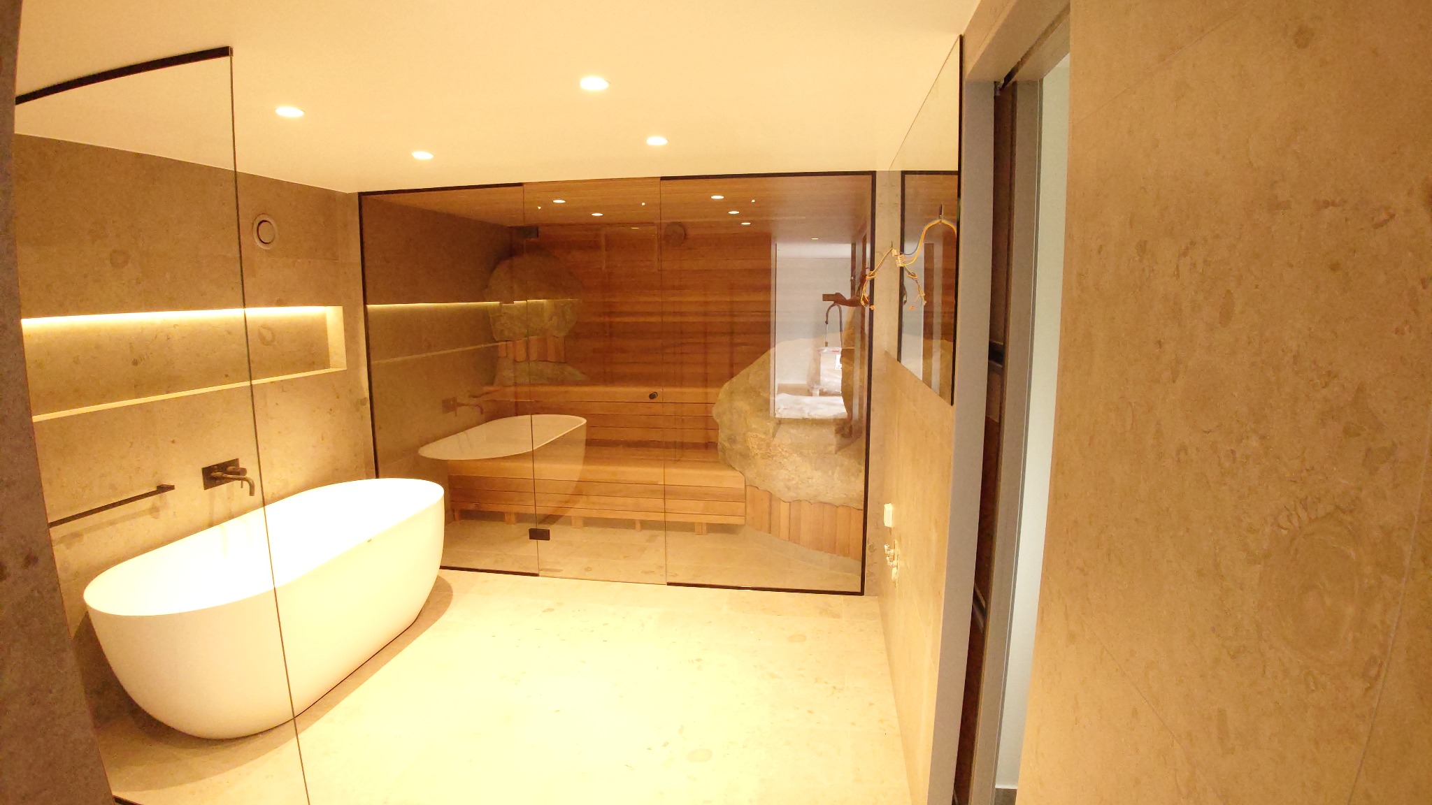 Nowoczesna łazienka z sauną oddzieloną szklaną ścianą, wolnostojąca biała wanna na tle kamiennej ściany i drewnianej sauny, beżowe płytki na ścianach i podłodze, oświetlenie punktowe.